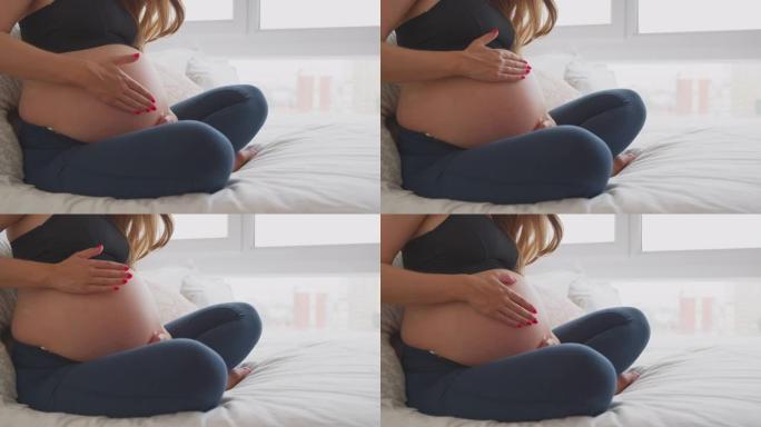 孕妇穿着健身服在家坐在床上摸肚子的特写
