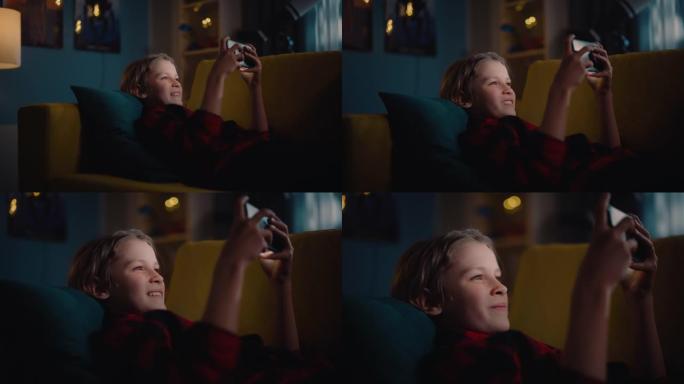 兴奋的小男孩躺在沙发上，在家里舒适的黑暗房间里用智能手机玩电子游戏。快乐成功的少年击败玩家并在在线比