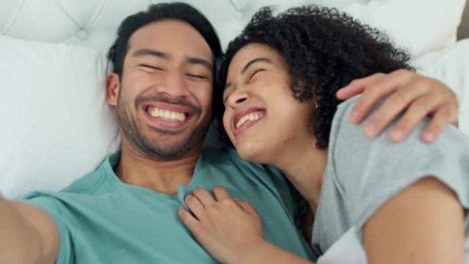 跨种族的夫妇在床上用手机自拍，在卧室放松的同时为社交媒体流媒体视频，在家里为幸福的婚姻微笑。漫画，爱