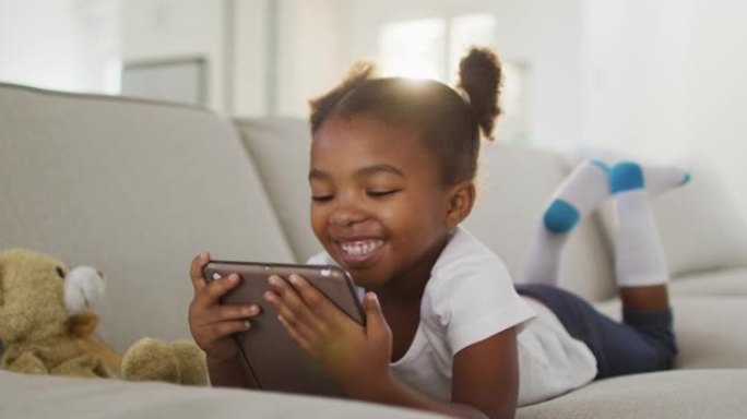快乐的非洲裔美国女孩躺在沙发上在家使用数字平板电脑