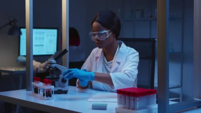 一位年轻的黑皮肤医生戴着防护眼镜，戴着手套和白色实验室外套，在实验室中标记了血液样本试管