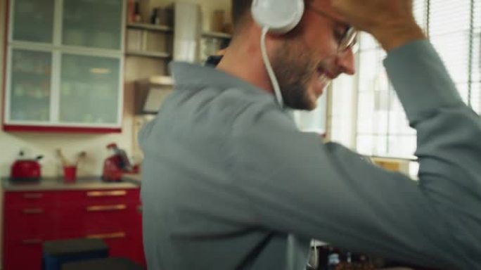 一个戴着白色耳机的年轻无忧无虑快乐男人的真实特写镜头很有趣，可以用智能手机听音乐，并在家里的厨房里疯