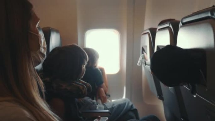 戴着医用面具的年轻女子和两个十几岁的孩子在飞行过程中看着飞机窗外的座位，看着令人惊叹的景色。