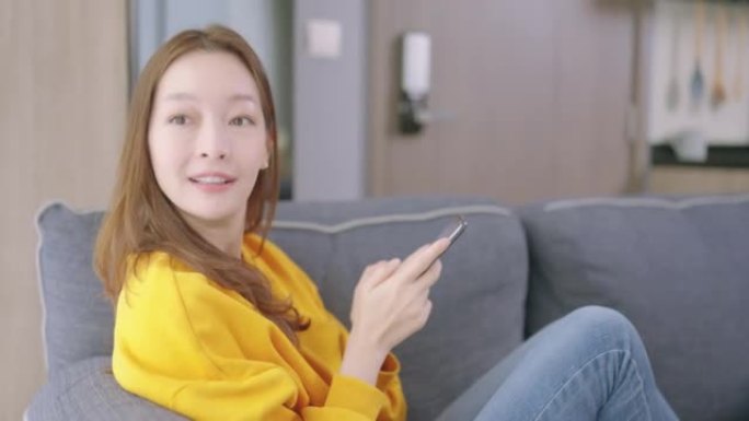 美丽的亚洲年轻女子年龄36岁，穿着五颜六色的黄色衬衫工作和手机网上购物，早上在家坐在沙发上放松。人和
