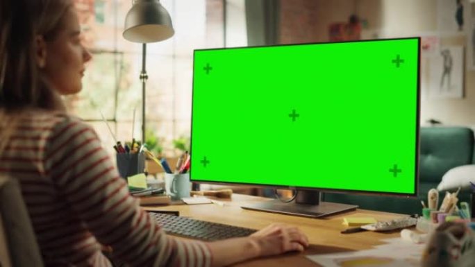 美丽的专业创意员工在她的个人计算机上工作，并带有绿色大屏幕模拟显示屏。她在凉爽的阁楼空间工作。学习和