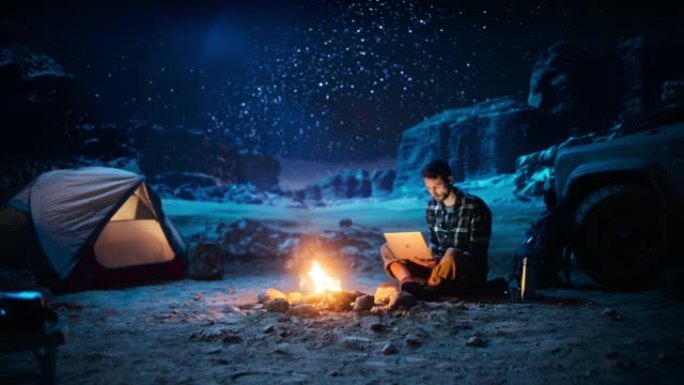坐在篝火旁的男性旅行者在峡谷中露营时使用笔记本电脑。在度假旅行中通过互联网进行数字远程工作，电子商务