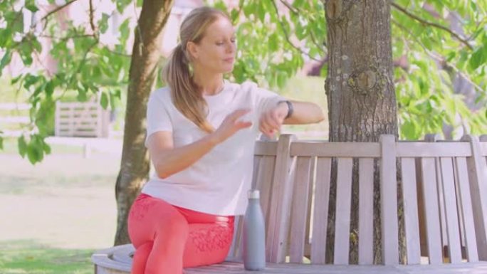 女人穿着健身服坐在树下的座位上检查智能手表上的活动监视器