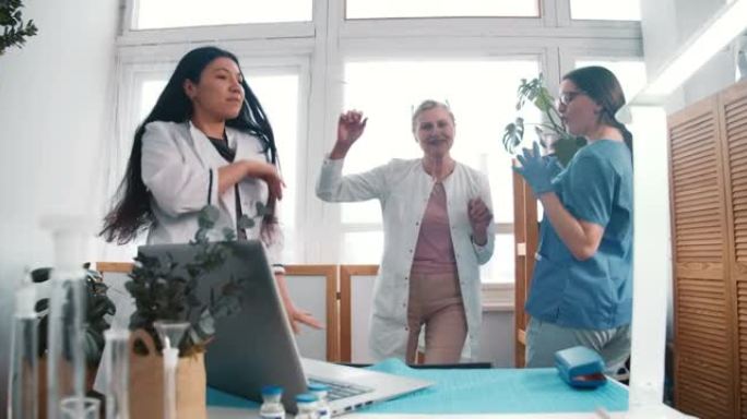抗击病毒大流行的结束。三位有趣的多民族快乐女医生在诊所实验室一起跳舞庆祝胜利。