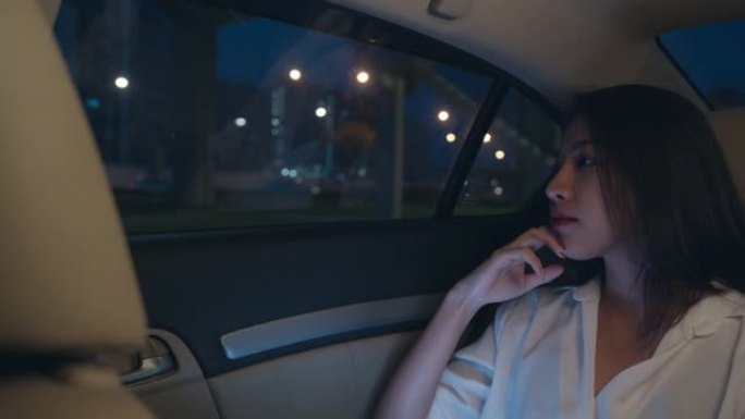 穿着时尚办公室服装的亚洲女商人晚上坐在城市现代的乘客后座上，看着窗外的车。