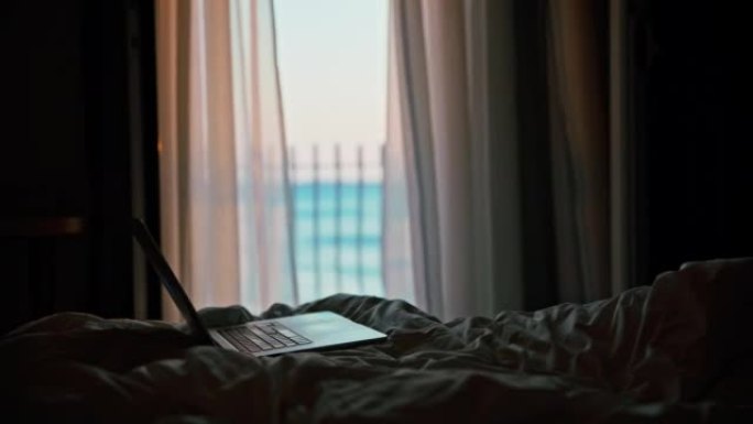 笔记本电脑站在酒店房间的床上，背景是阳台和海，透明的窗帘