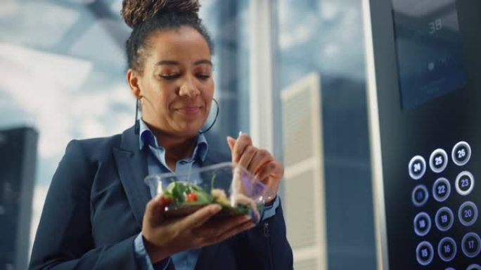 自信的黑人女性乘坐玻璃电梯到现代商务中心的办公室，吃健康的蔬菜沙拉。快乐的经理为工作中富有成效的一天