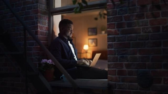 年轻英俊的黑人在家工作，在舒适时尚阁楼公寓的窗台上的笔记本电脑上工作。富有创造力的男性微笑，检查社交
