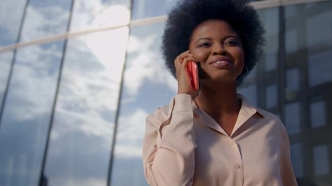 一位皮肤黝黑的年轻卷发妇女在粉红色的iPhone上微笑着说话，经过商务中心