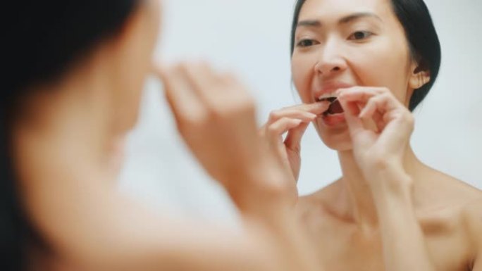 美丽的亚洲女人的肖像用牙线清洁她的天然洁白的牙齿，微笑在浴室的镜子。快乐的女性享受早晨的例行公事。牙