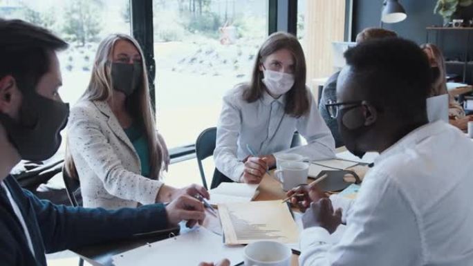 有疫情安全措施的工作场所。年轻的多民族商业同事在办公室会议上工作，戴着口罩。