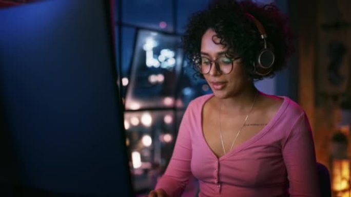 晚上，在时尚的阁楼公寓里，戴着耳机的美丽多民族黑人妇女使用电脑。有创意的女性微笑，在社交媒体上浏览视