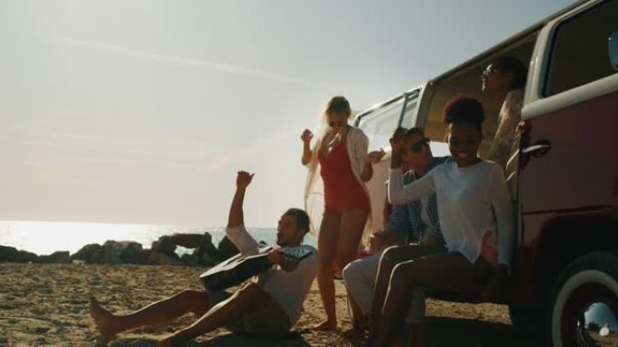 一群年轻的多民族朋友的慢动作来到海边的海滩上，玩得很开心，弹吉他，唱歌和跳舞，阳光明媚。