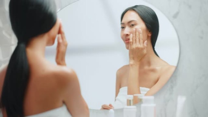 美丽的亚洲女人的肖像轻轻地涂抹面霜面膜，用性感的触摸，看着浴室的镜子。年轻女性让她的皮肤柔软，清洁与
