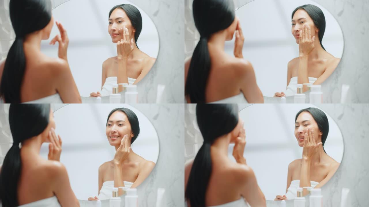 美丽的亚洲女人的肖像轻轻地涂抹面霜面膜，用性感的触摸，看着浴室的镜子。年轻女性让她的皮肤柔软，清洁与