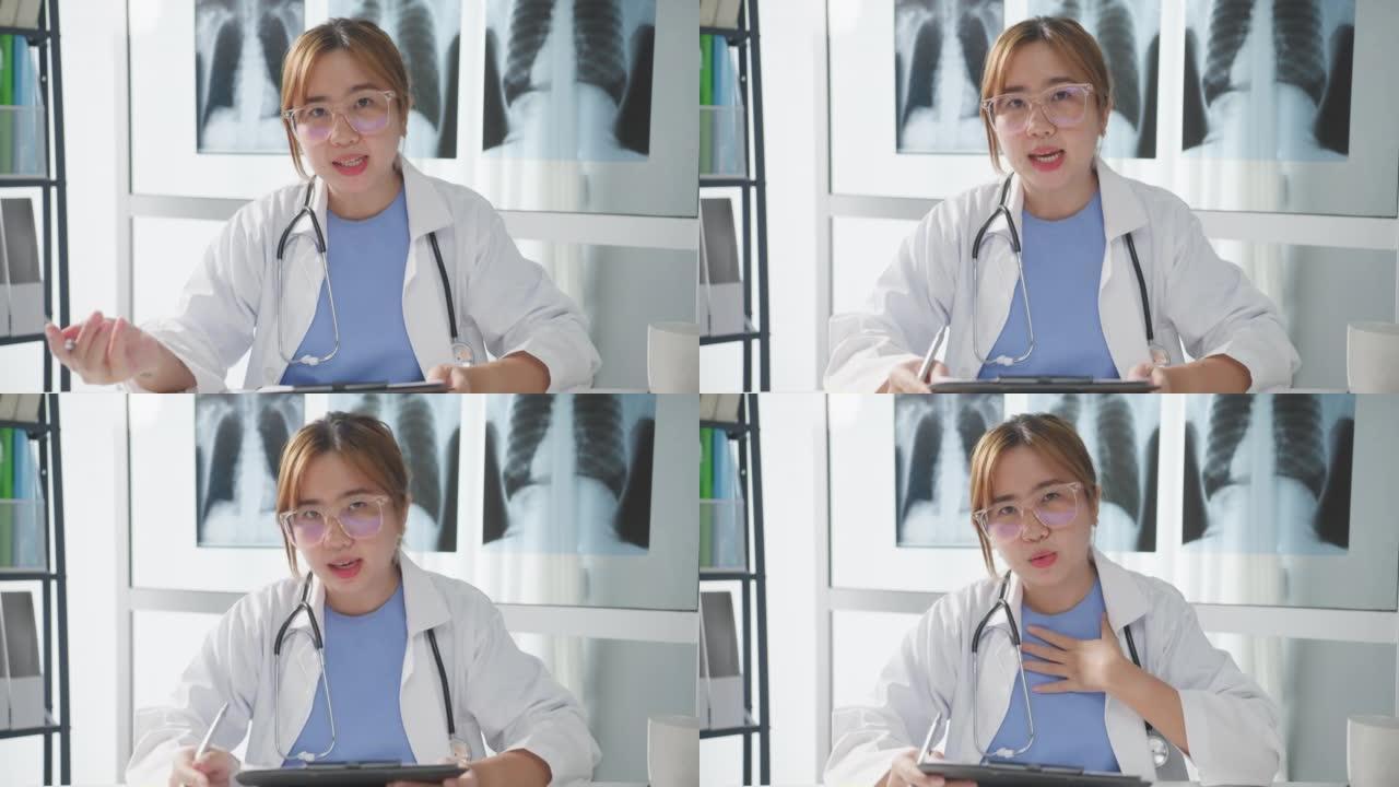 年轻的亚洲女士医生穿着白色医疗制服，用听诊器用电脑笔记本电脑与病人交谈视频电话会议，看着医院的摄像头