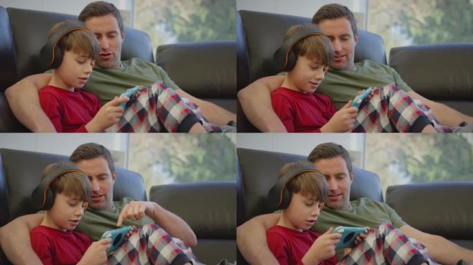 父亲和儿子戴着无线耳机在手持设备上玩电脑游戏-慢动作拍摄