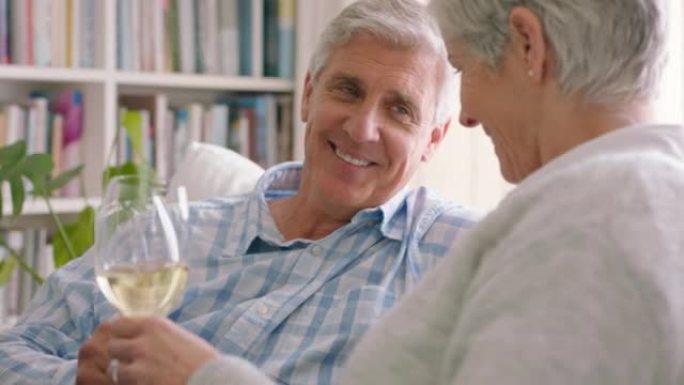 老年夫妇在他们现代房子的客厅沙发上喝香槟或葡萄酒。退休时快乐、积极、大笑的老年人，在家里的沙发上享受