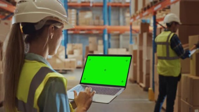 专业女工戴着安全帽拿着带绿色色度键屏幕的笔记本电脑，在摆满了货架的零售仓库中，在风景模式下摆满了送货