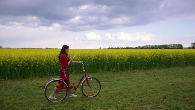 慢动作，跟踪一个穿着红色连衣裙的黑发女人的镜头，看着她的手机，她骑着自行车穿过一片充满金鱼草的草地