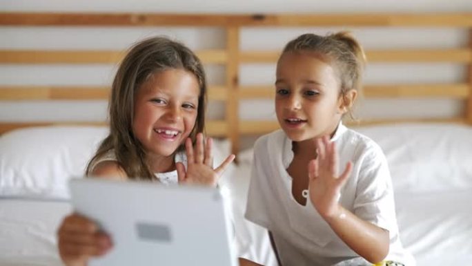 两个快乐的小姐妹的真实照片很开心，坐在床上用平板电脑给父母或亲戚做自拍或技术视频通话