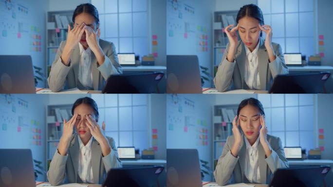年轻的亚洲女商人坐在桌子上，用笔记本电脑和平板电脑摩擦眼睛，晚上在办公室因劳累而感到疼痛和疲倦。