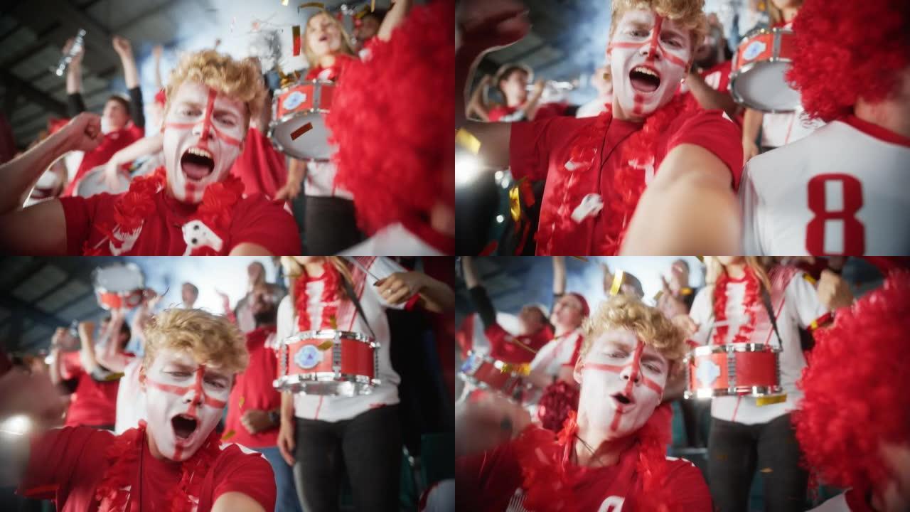 体育场体育赛事: 画脸男子的自拍肖像拿着智能手机开枪，为红队获胜欢呼，尖叫，与人群一起庆祝冠军胜利