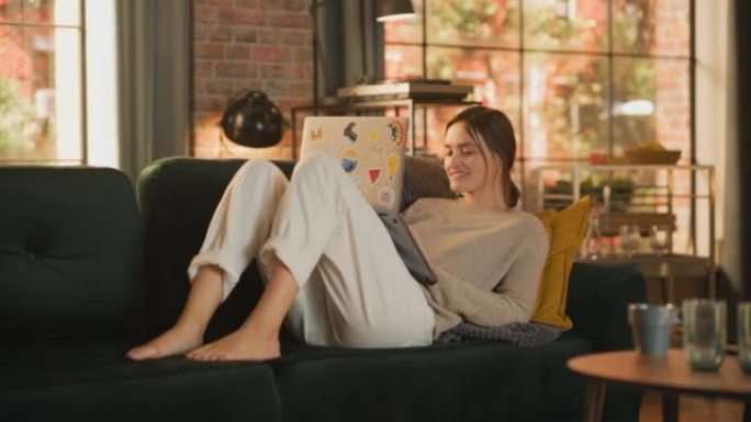 年轻漂亮的女人躺在沙发上，在阳光明媚的日子里在时尚的阁楼公寓里使用笔记本电脑。富有创造力的女性微笑，