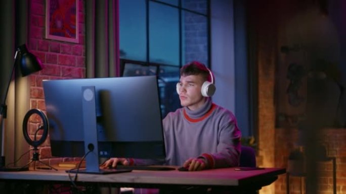 戴着耳机的年轻帅哥晚上在时尚的阁楼公寓里用电脑。有创造力的男性在家工作，浏览互联网和社交媒体。从大窗