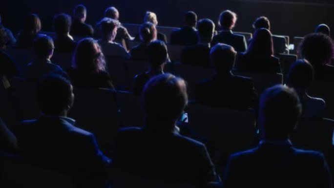 一群商界人士在黑暗的会议厅里观看创新的鼓舞人心的主题演讲。商业技术峰会礼堂会议室挤满了企业代表。后面