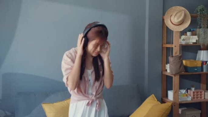 快乐无忧无虑的年轻亚洲女孩戴着耳机听音乐独自在家里的客厅跳舞。隔离社会距离冠状病毒大流行概念。享受自