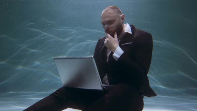 创造性的职业概念。年轻快乐的商人穿着西装思考，用笔记本电脑在水下慢动作下工作。