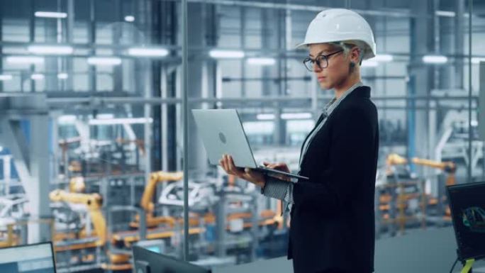 女工程师戴着白色安全帽，使用笔记本电脑，在汽车装配厂的办公室里望着外面。在技术设施中从事车辆设计的工