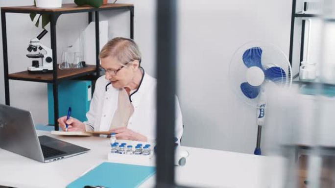 穿着实验室外套的友好的高级高加索医生妇女，眼镜微笑使用笔记本电脑网络摄像头聊天咨询远程患者