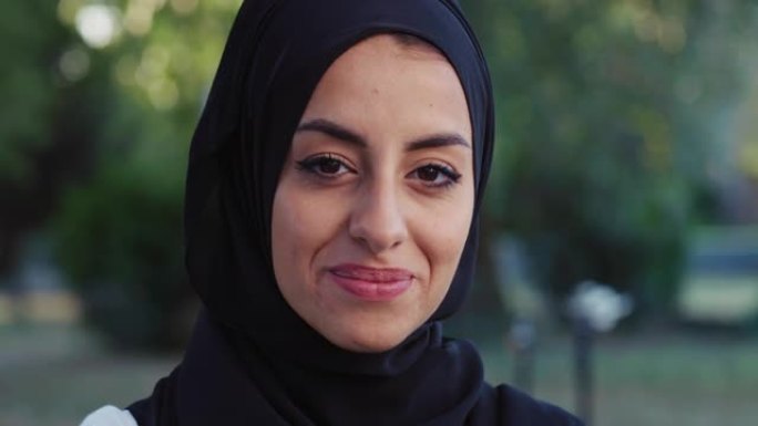 美丽的年轻阿拉伯女子微笑着绿色背景的肖像。带着头巾的快乐穆斯林女性运动员在清晨锻炼时在公园享受新鲜空