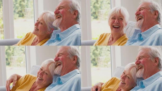 退休的高级夫妇坐在家里休息室的沙发上，一起观看摄像机电视和慢动作大笑