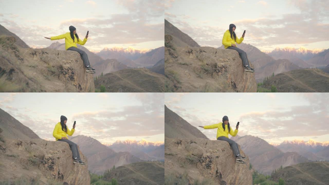 快乐的自由旅行者穿着黄色冬季夹克的年轻女性坐在岩石上通过智能手机通话视频给她的朋友，并在冬季的山顶上