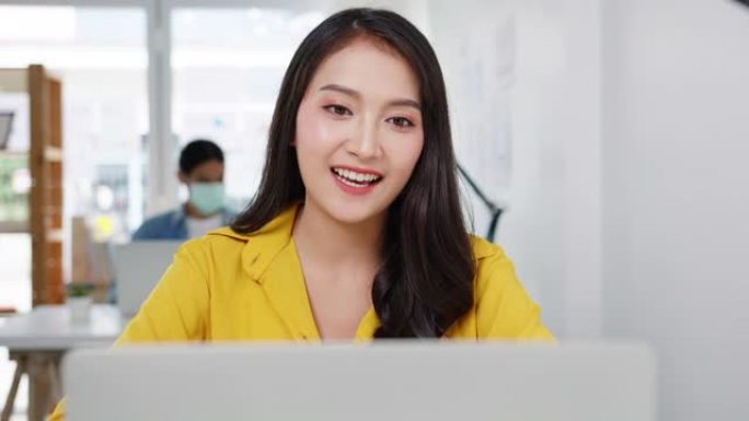 亚洲女商人在新的正常情况下进行社交距离，以预防病毒，同时在办公室工作时使用笔记本电脑向同事介绍有关视
