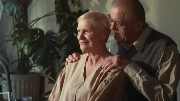 团结和爱。快乐浪漫的70年代高级丈夫拥抱美丽的白发妻子，在舒适的家庭厨房交谈。