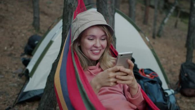 快乐的年轻女子使用智能手机发短信，在森林的吊床上放松的信息上大笑