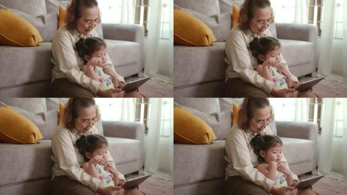 可爱的孙女坐在微笑和快乐的祖母在家里的数字平板电脑上观看卡通直播。老少都要学习新技能