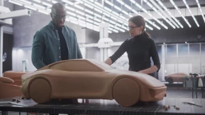 年轻的女学徒汽车建模师与黑人高级汽车设计师一起为创建现代电动汽车的3D聚合物粘土模型提供建议。在创意