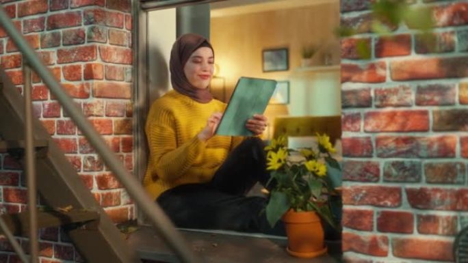 穆斯林女孩戴着头巾，坐在布鲁克林风格的褐砂石房子的窗台上，使用平板电脑。年轻女性浏览互联网。从房子外