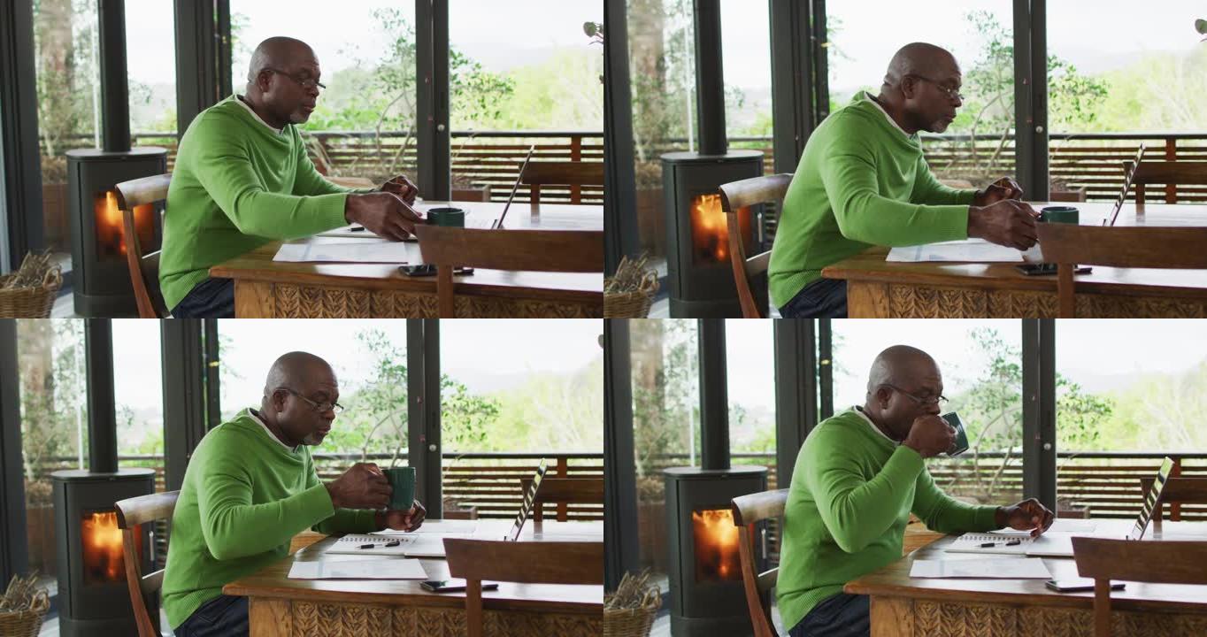 非裔美国高级男子坐在餐桌上工作，使用笔记本电脑并喝咖啡