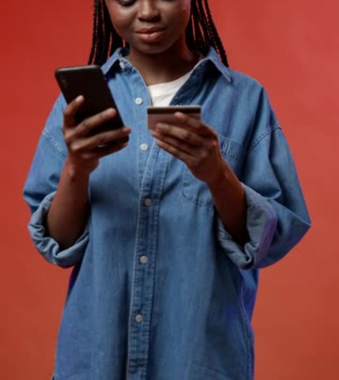 一个开朗迷人的深色皮肤博客作者，长长的非洲辫子站在红色背景下，她在网上商店用信用卡付款
