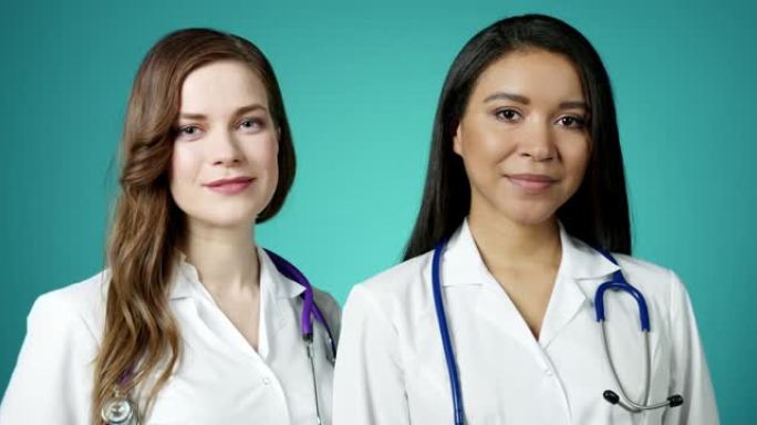 美丽年轻快乐的医务女孩对着镜头微笑。两位成功自信的现代女医生穿着白大褂慢动作摆姿势。女性医学工作者。